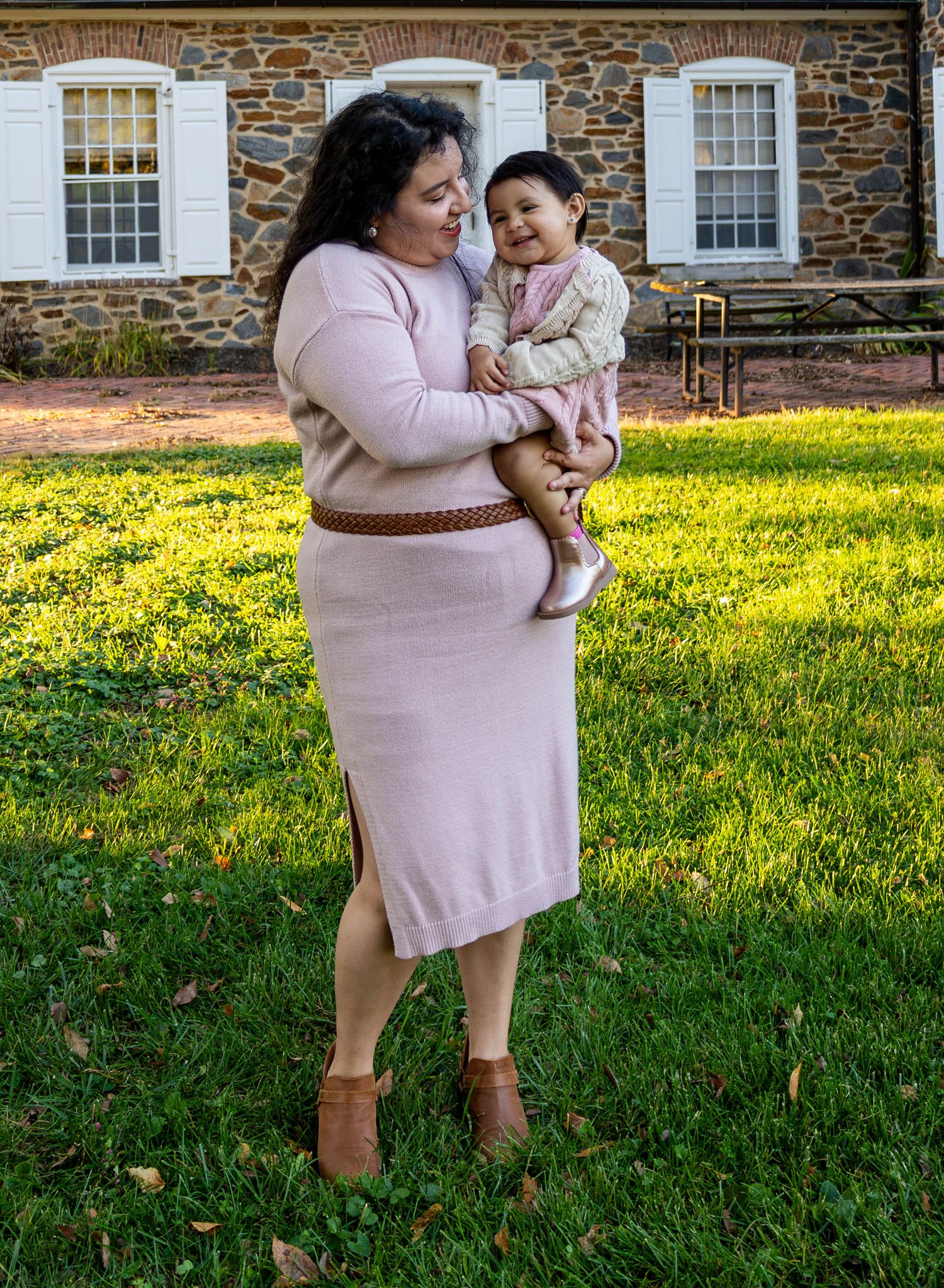 A woman in a pink dress holds a smiling baby outdoors on green grass near a stone building.