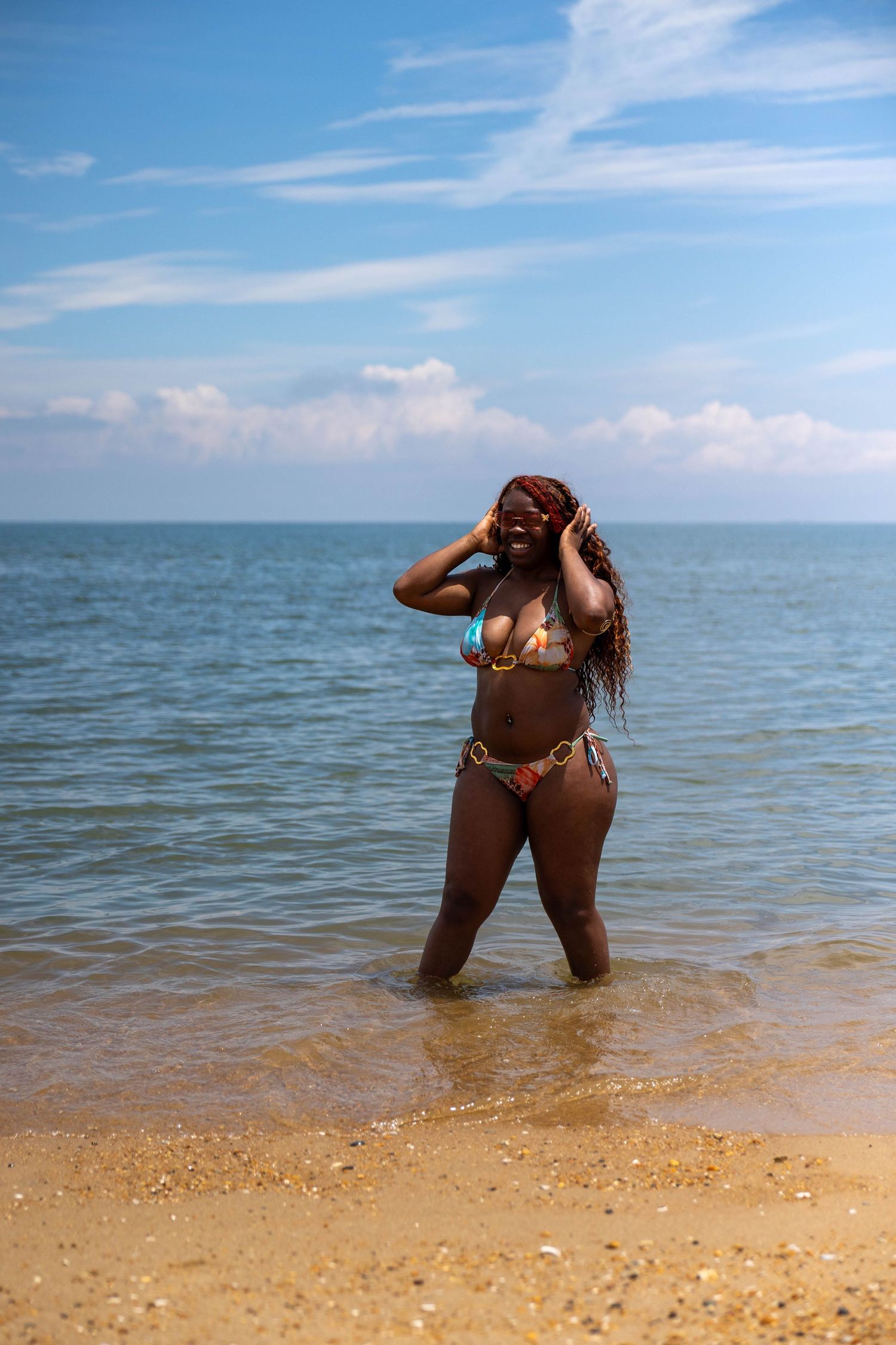 A curvy Black woman in a colorful bikini stands in shallow ocean water on a sunny beach.