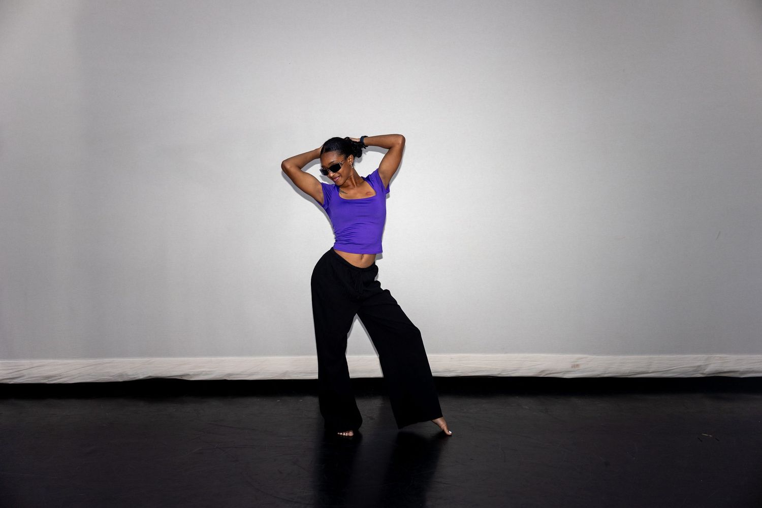 A dancer in a purple crop top and black wide-leg pants poses gracefully with arms raised on a black stage floor.