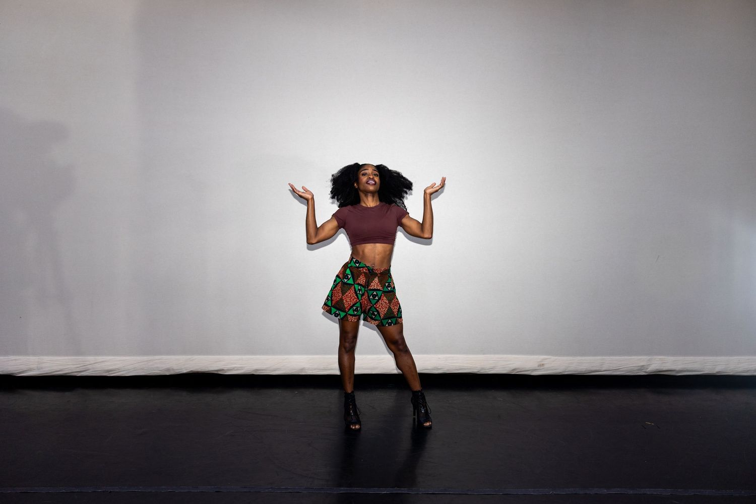 Black woman in African print skirt dancing expressively with arms raised on a dark stage floor.