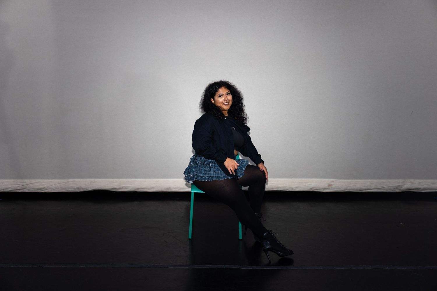 A woman sits thoughtfully on a green stool in a dark jacket and skirt in a minimalist studio setting.