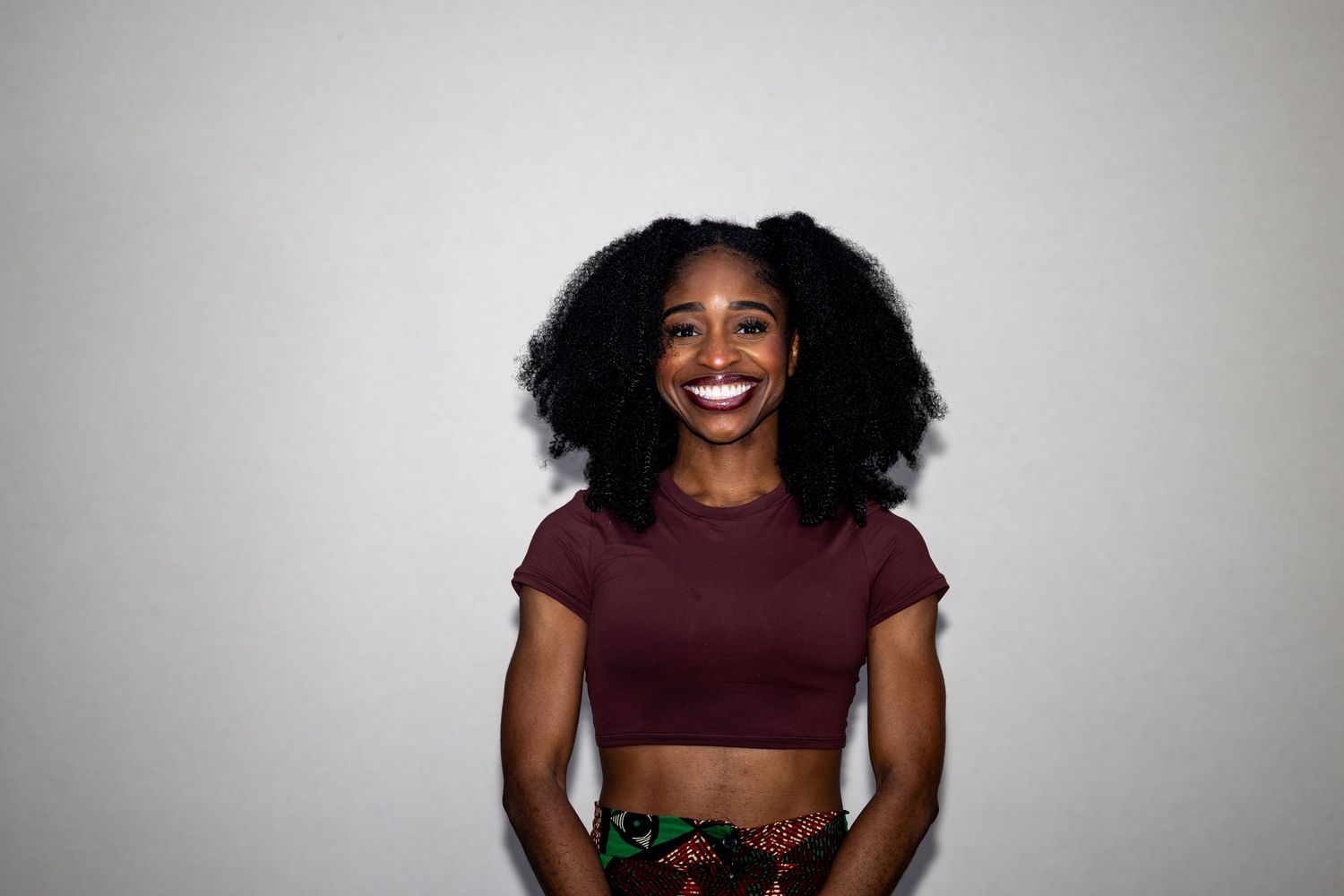 Smiling young Black woman with natural curly hair wearing a maroon crop top and colorful printed skirt against a white background.