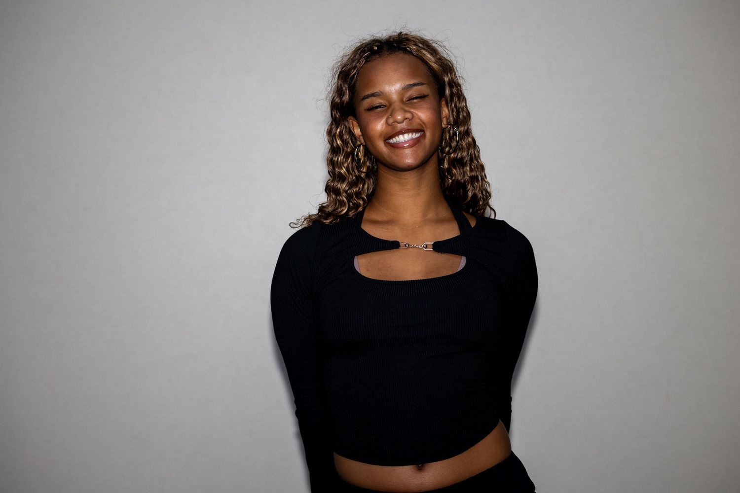 A smiling young woman with curly highlighted hair wears a black cutout long-sleeve crop top against a gray background.
