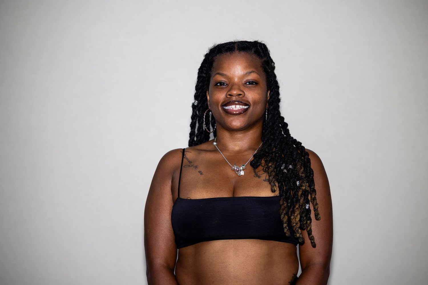 Smiling Black woman with braids wearing a black crop top poses against a light gray background.