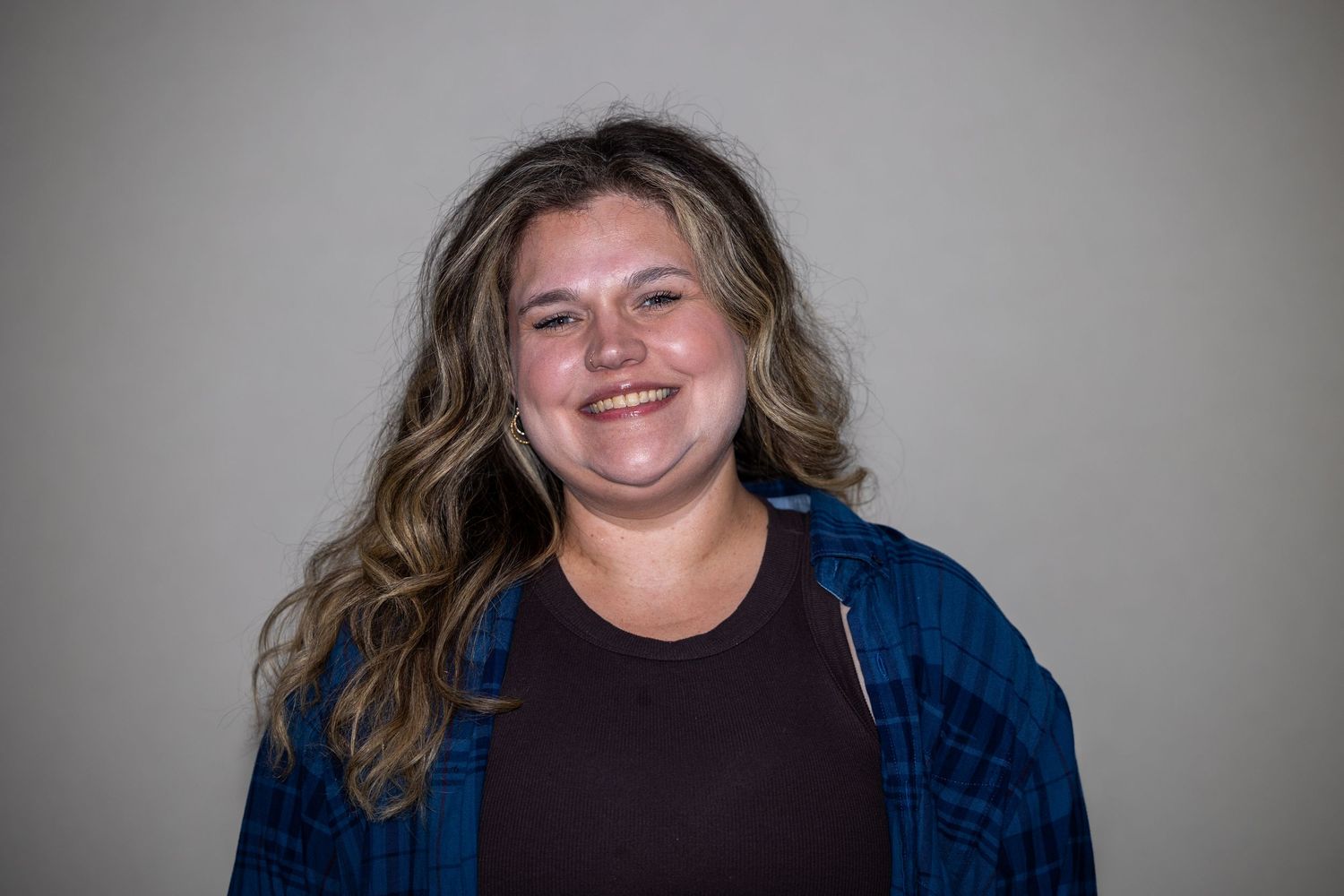 Smiling young woman with wavy blonde hair wearing a blue flannel shirt and dark top against a gray background.