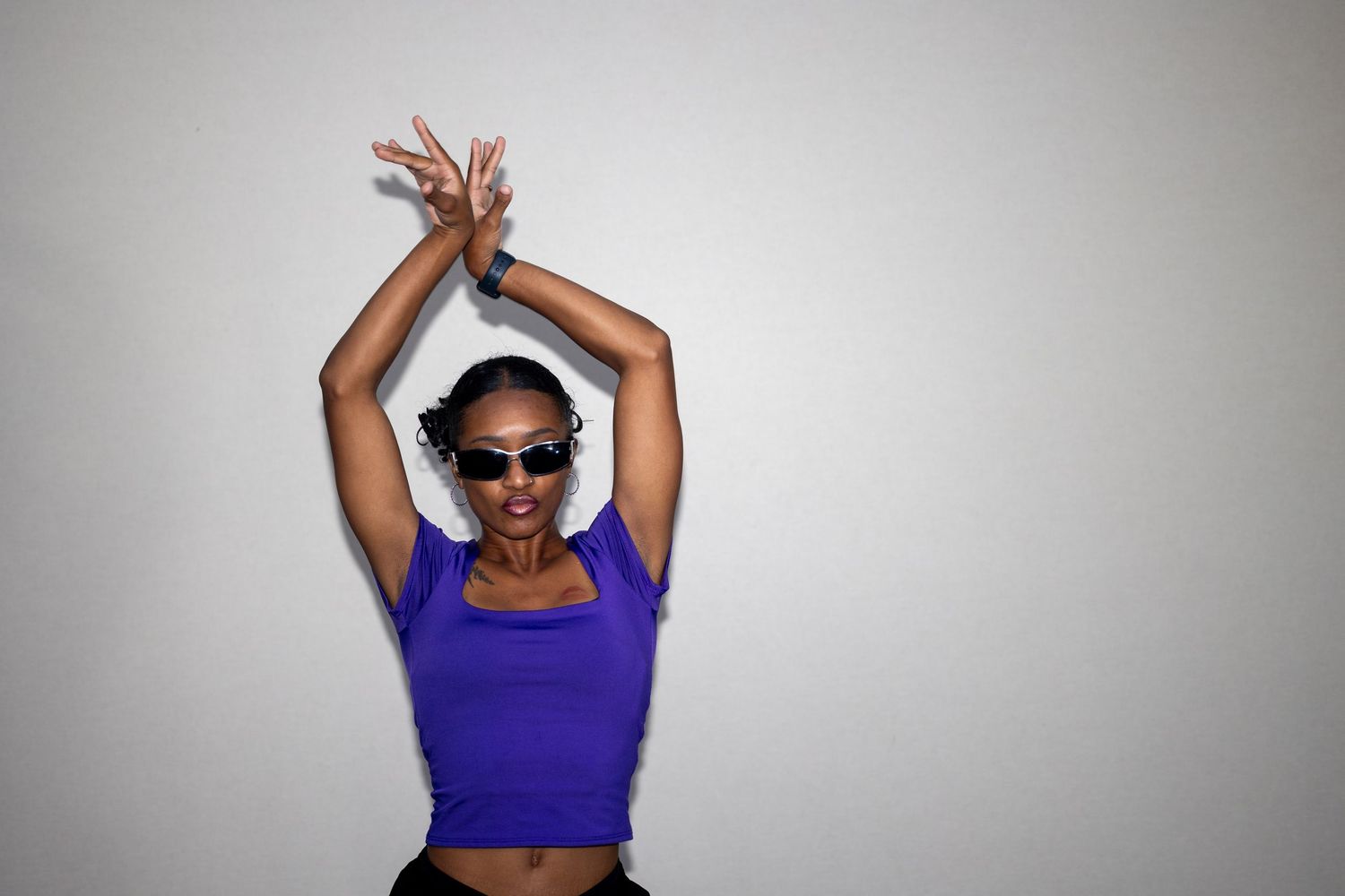 Woman in purple crop top and sunglasses poses expressively with raised arms against white background.