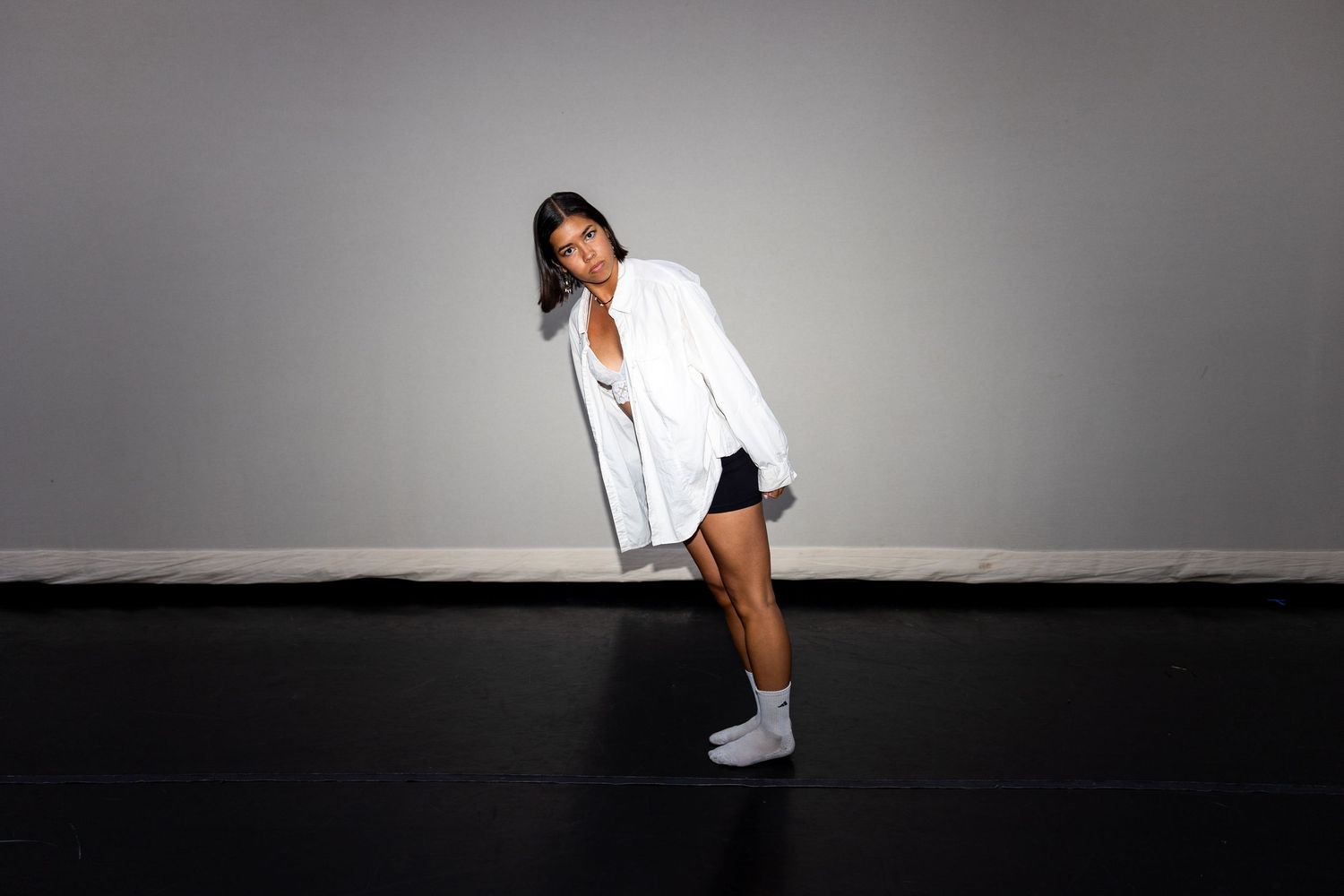 A dancer in a white blazer and black shorts leans gracefully in a studio setting.