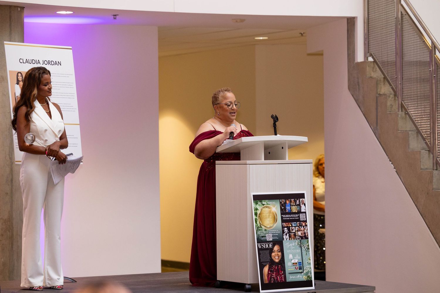 Woman in burgundy dress speaks at podium during Claudia Jordan event while woman in white outfit stands nearby.
