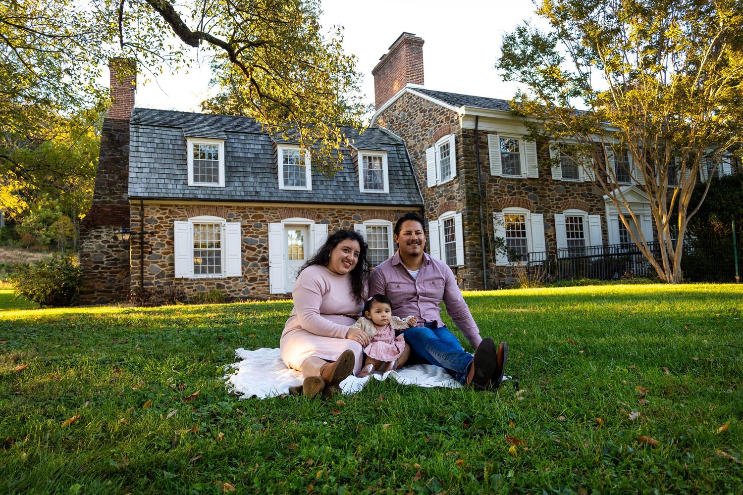 Family of three sitting on green grass in front of a large stone colonial home during golden hour.