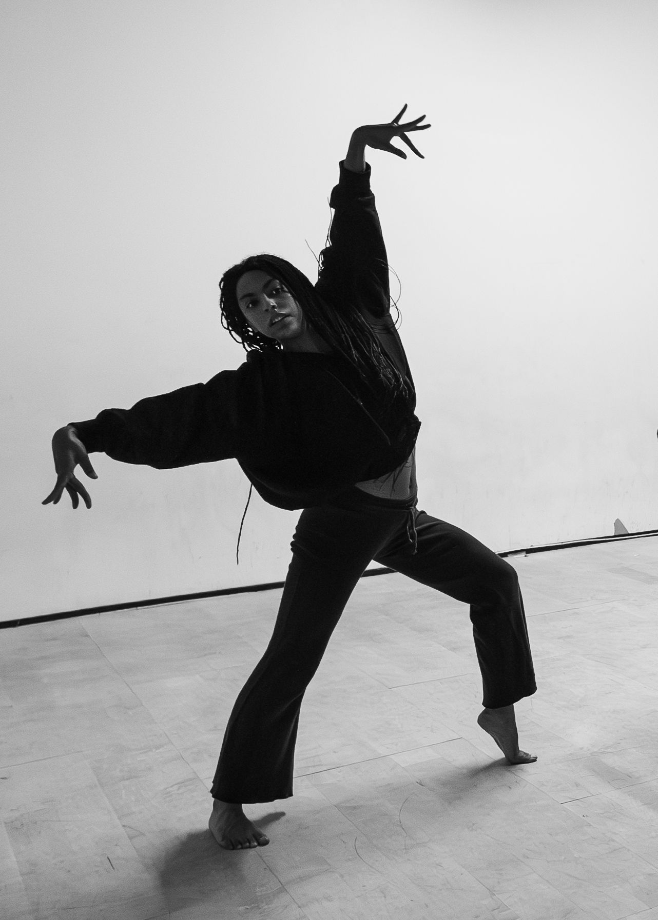 Black and white photo of a dancer in dramatic pose wearing all black, one arm raised, in a studio.