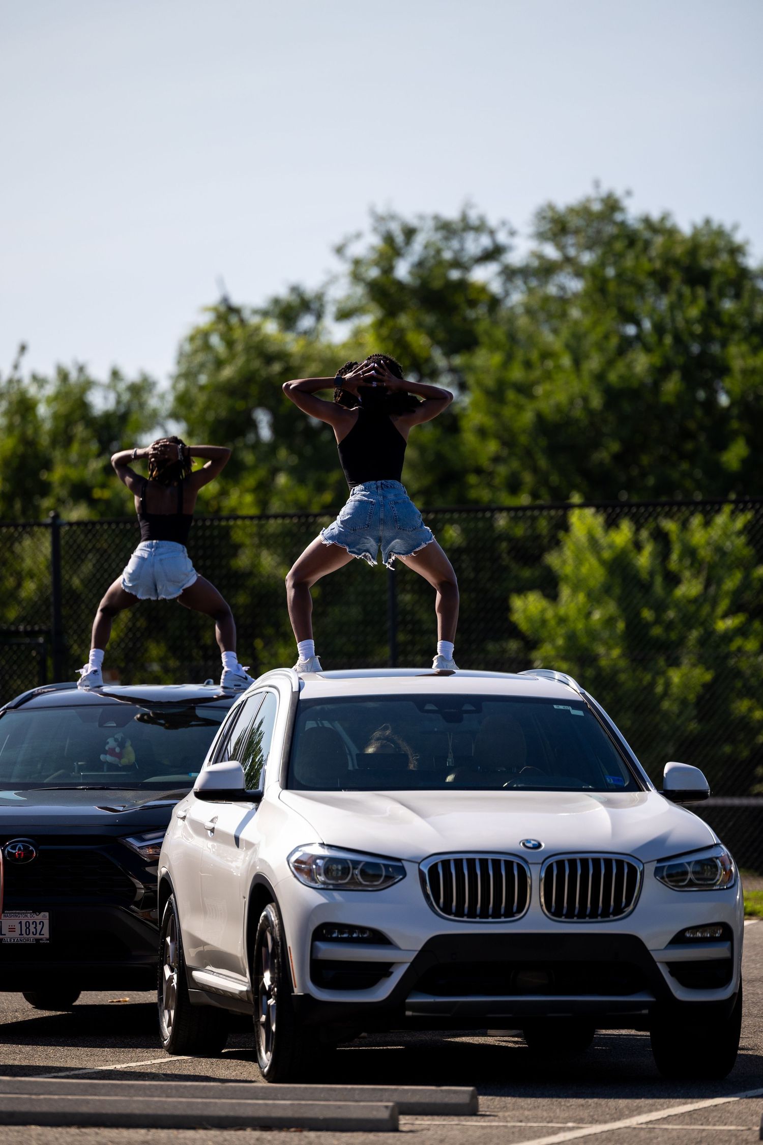 Two people standing on top of a white BMW SUV in a parking lot, posing with hands behind their heads.