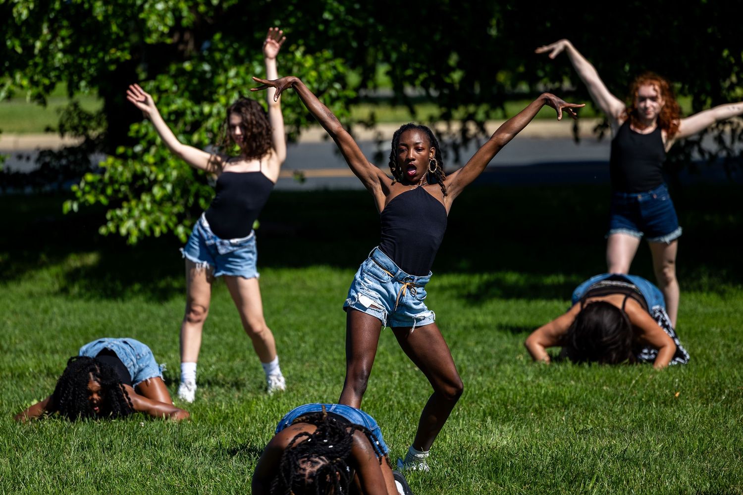 Young women dancing outdoors on green grass in denim shorts and black tops, performing expressive choreography in sunlight.