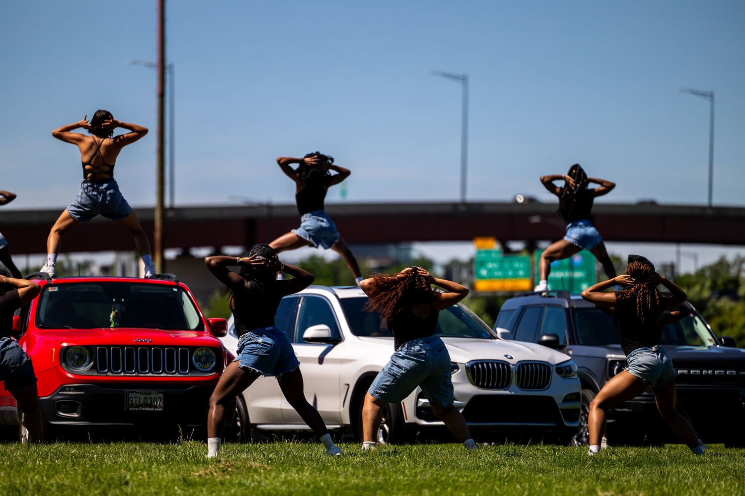 Group of people posing on and around cars, including a Jeep and BMW, on a sunny day near a highway overpass.