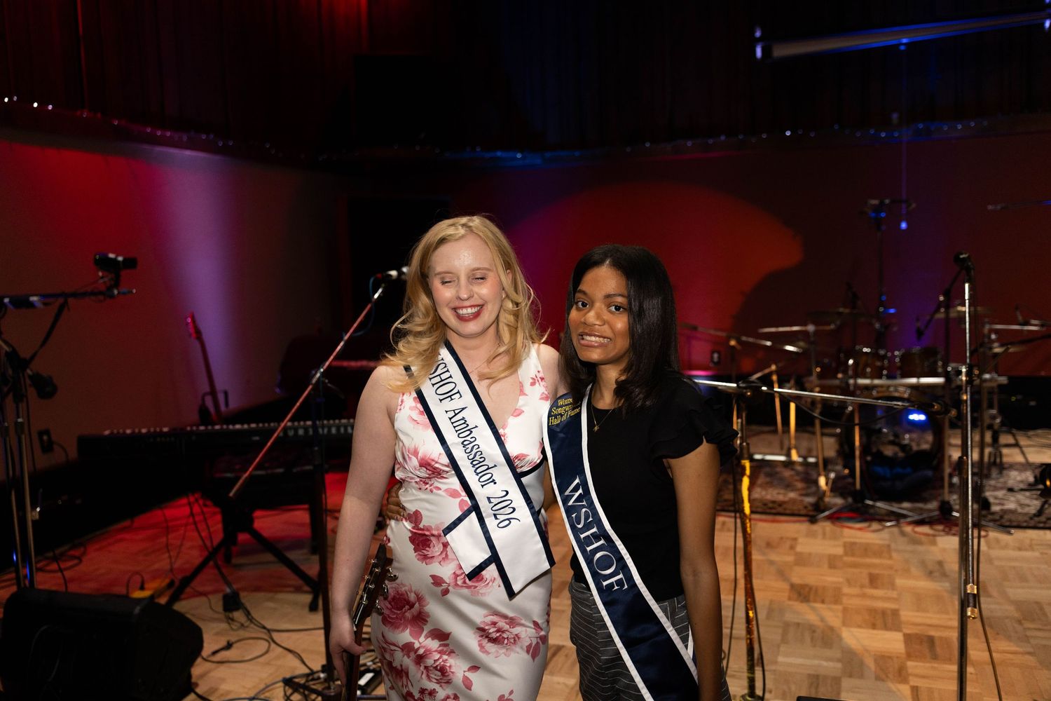 Two women wearing pageant sashes pose together smiling in a dimly lit venue with musical equipment in the background.
