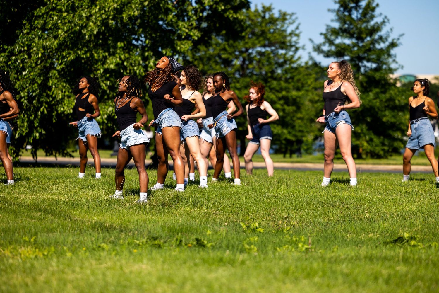 Group of young women dancing outdoors on green grass in a sunny park, wearing matching blue shorts and black tops.