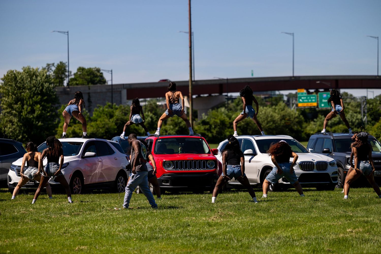 People dancing and jumping on cars parked on a grassy area near a highway on a sunny day.