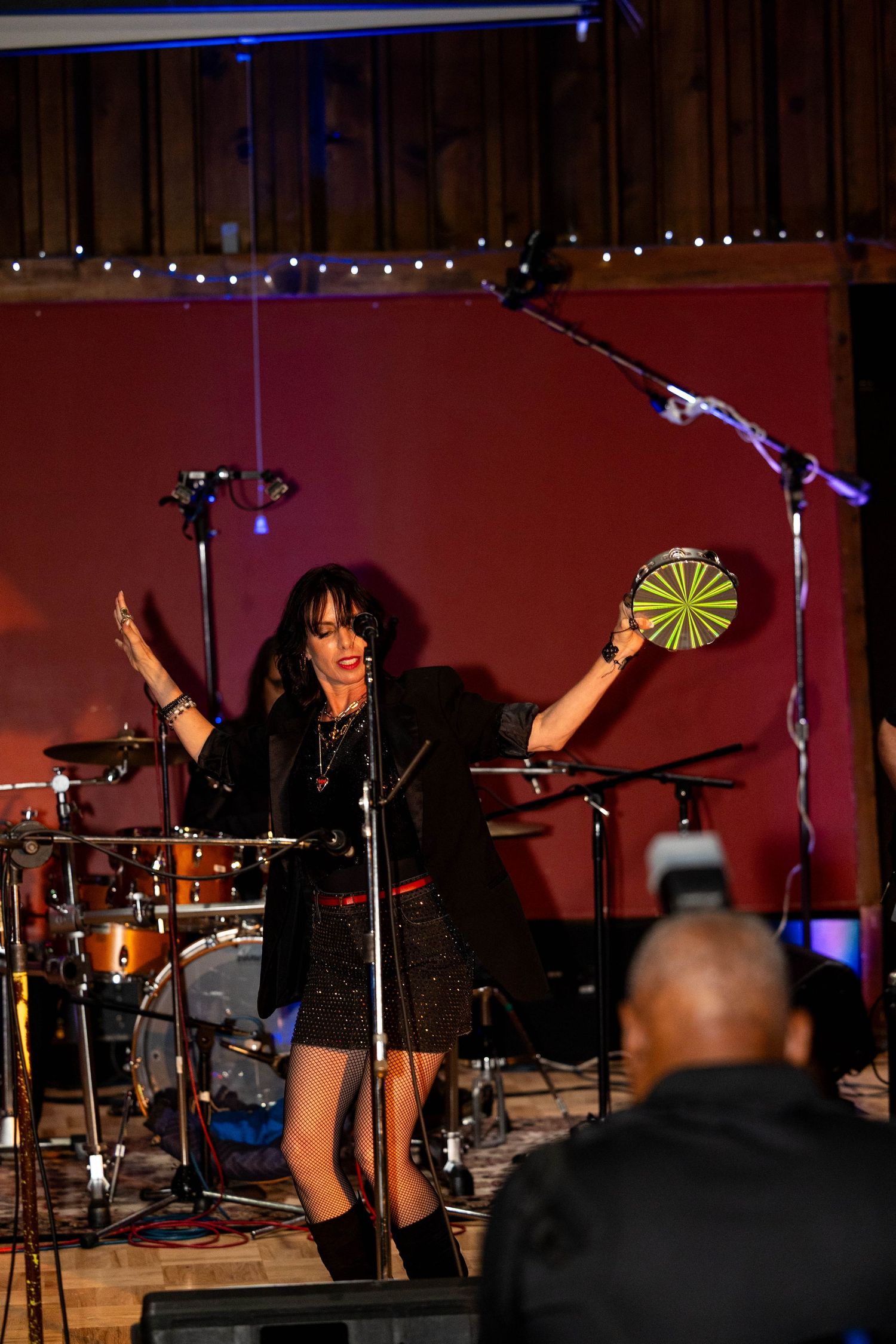 Female singer performing on stage holding a tambourine, with drums and microphone stand visible in a red-lit venue.