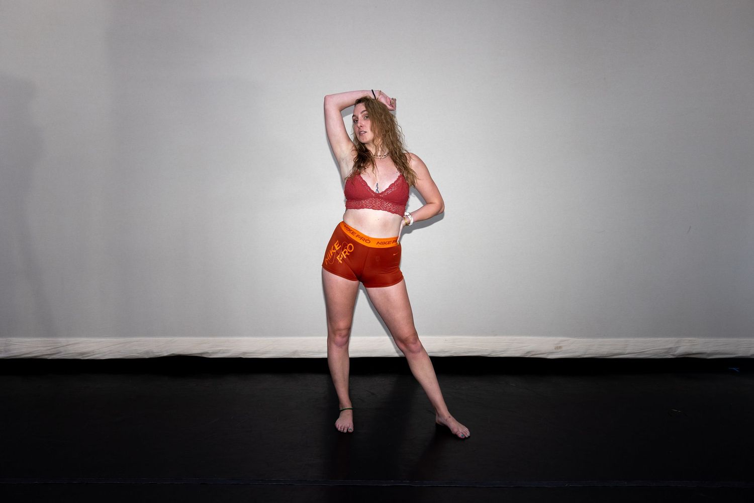 Female dancer in red and orange sports bra and shorts striking a pose in a white studio.