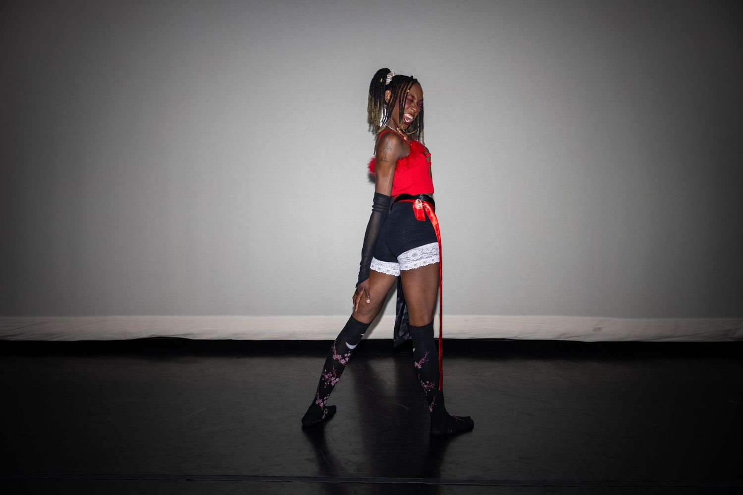 Female dancer in red top, black shorts, and knee-high boots performing on a dark stage.