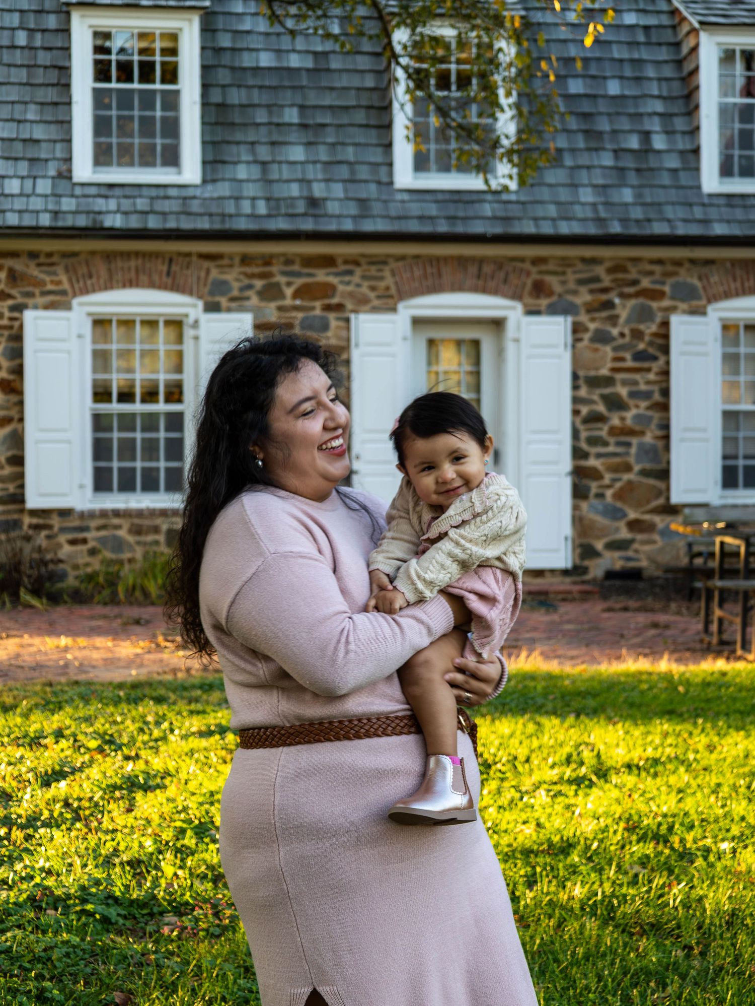 Smiling mother holding happy baby in front of stone colonial house on sunny day.