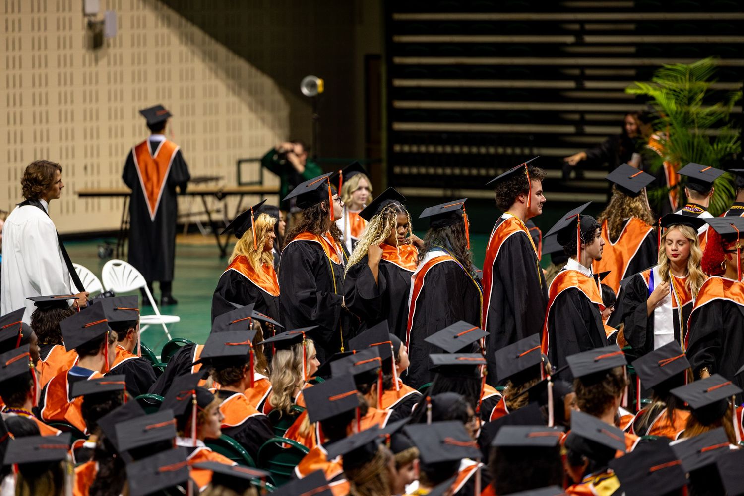 Graduates in black and orange caps and gowns gather at a commencement ceremony inside a large venue.