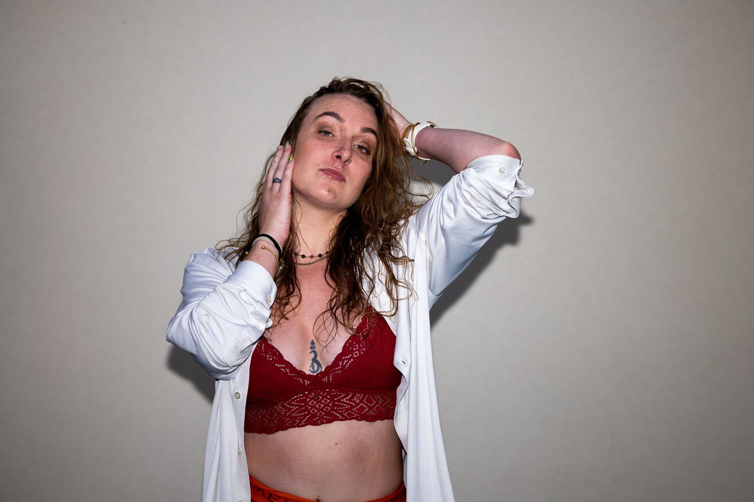 Young woman with wavy hair wearing a red lace bralette and open white shirt posing against a gray wall.
