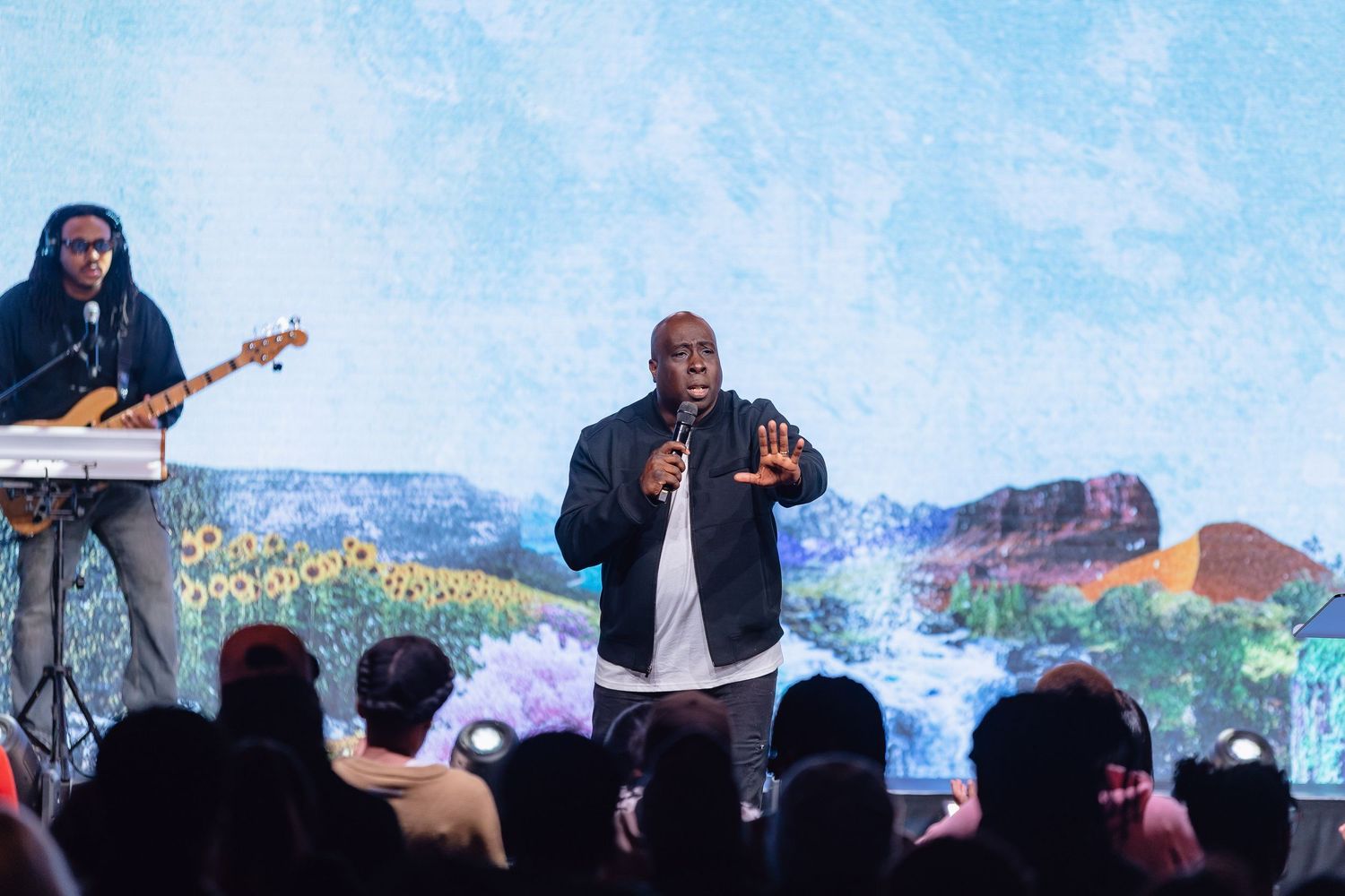 A performer gestures energetically on stage during a live musical performance with a colorful backdrop.