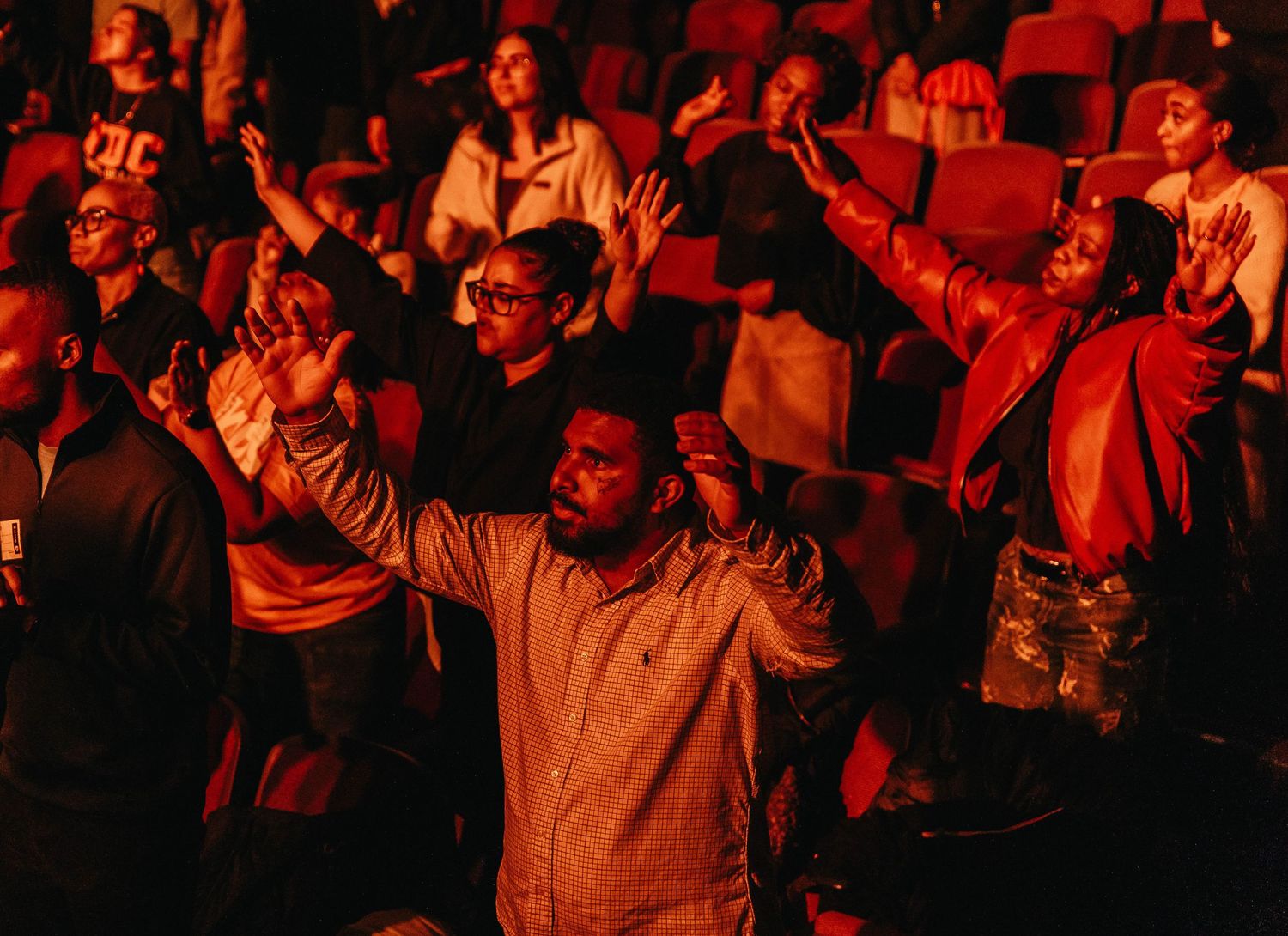 A large crowd of spectators in a dark venue illuminated by red lighting raises their hands in excitement.