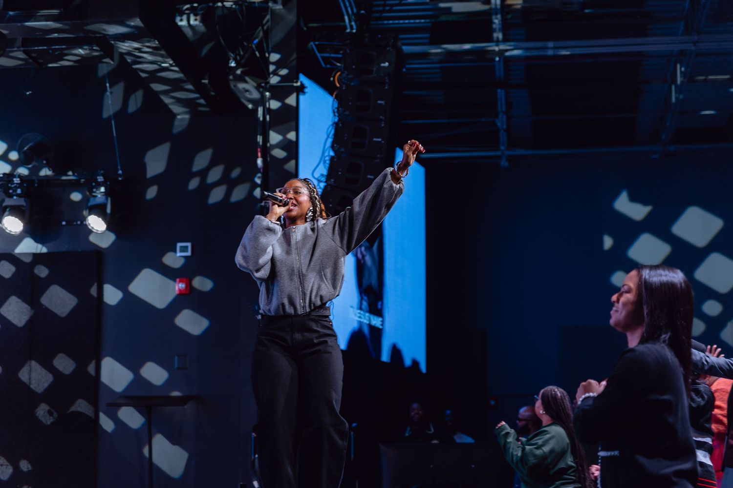 Concert crowd cheers with hands raised while watching a performer on stage in a dark venue with blue lighting.