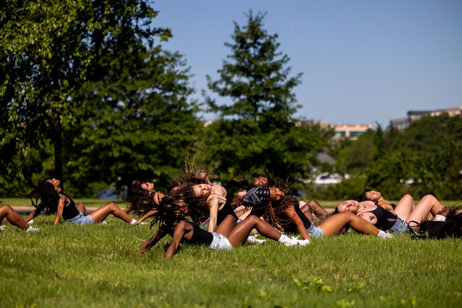 Group of young women relaxing on green grass in a sunny park with trees and city buildings in background.