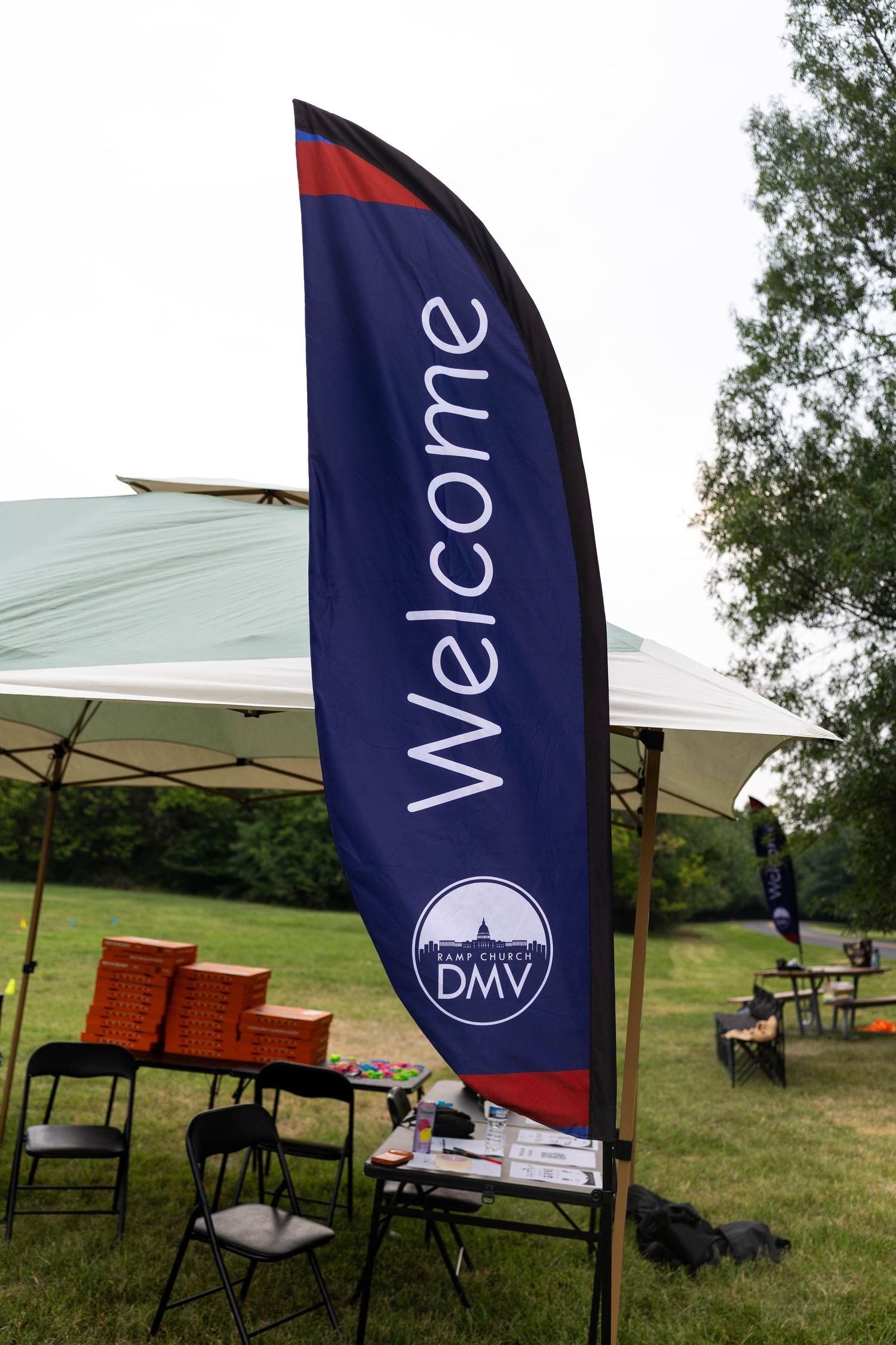 A navy blue Welcome feather flag for DMV Deaf Church stands at an outdoor event with a white tent and folding tables.