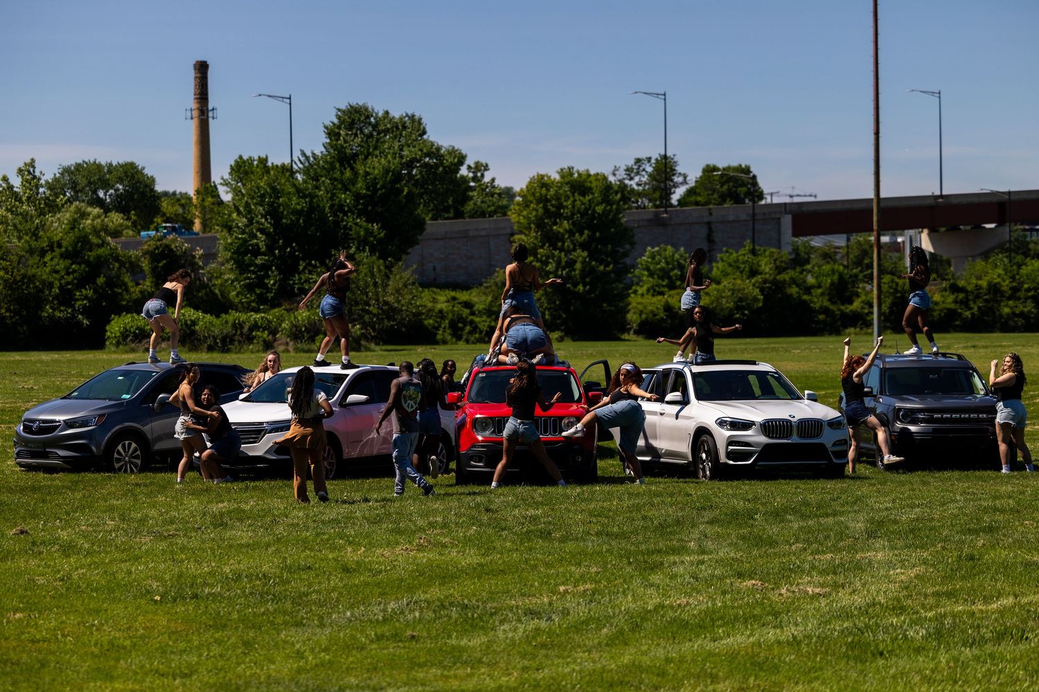 Cheerleaders posing with multiple SUVs and cars on a green field near an industrial area on a sunny day.