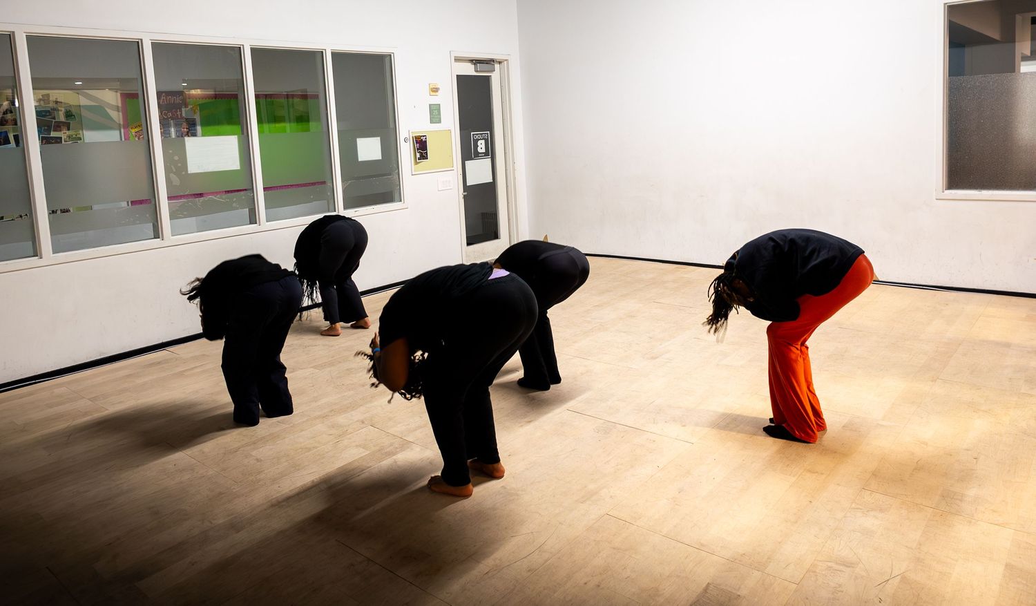 Three dancers in black bowing forward in a wooden-floored studio with white walls and green-framed windows.
