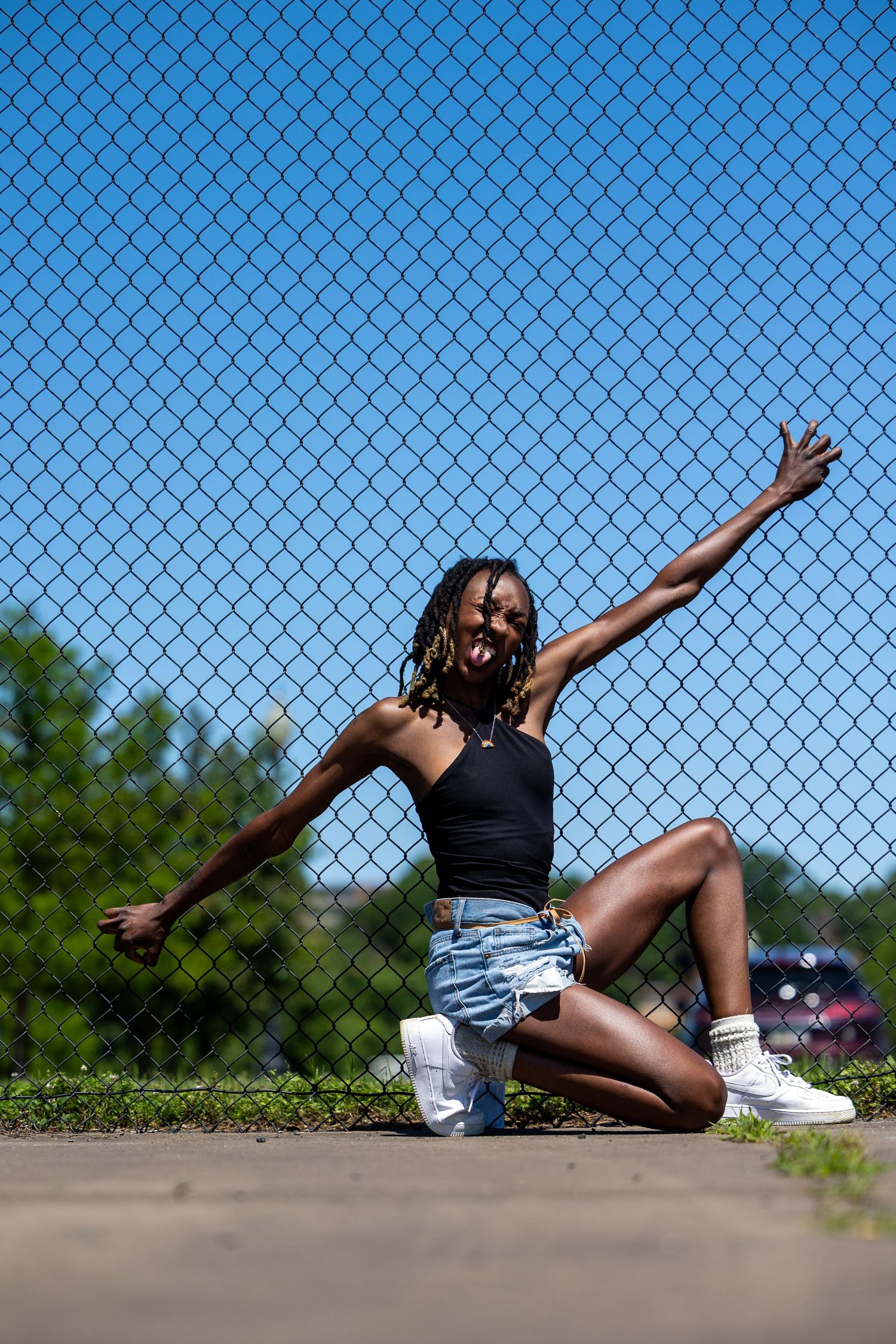 Young Black woman in denim shorts and crop top poses energetically against chain-link fence on sunny day.