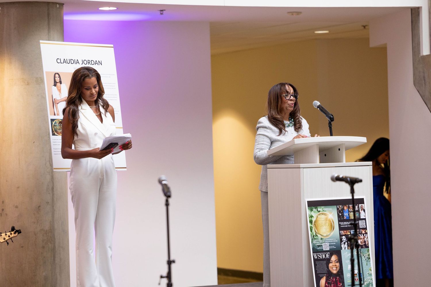 Two women in white outfits speak at a professional event, one at a podium and one holding papers on stage.