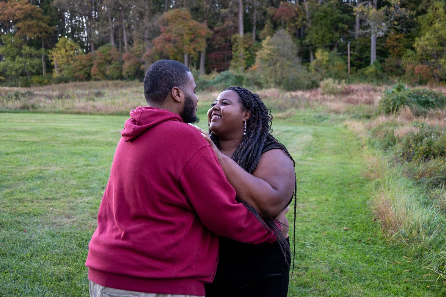 A joyful Black couple laughs together outdoors in a lush green field surrounded by autumn trees.