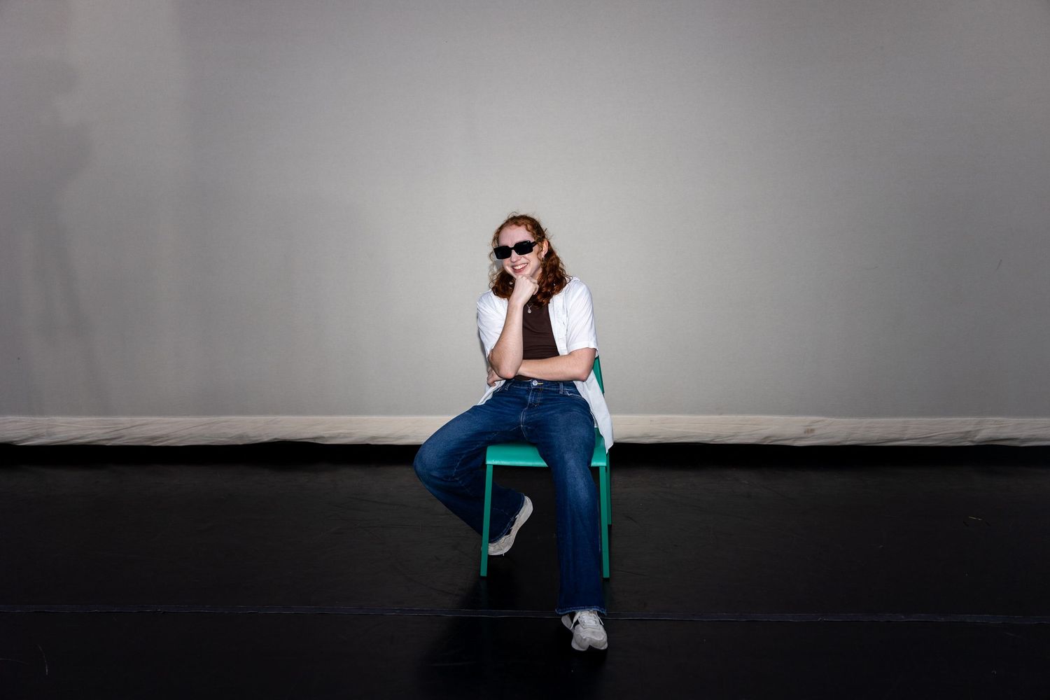 A smiling woman in jeans and white shirt sits relaxed on a green chair wearing stylish sunglasses.