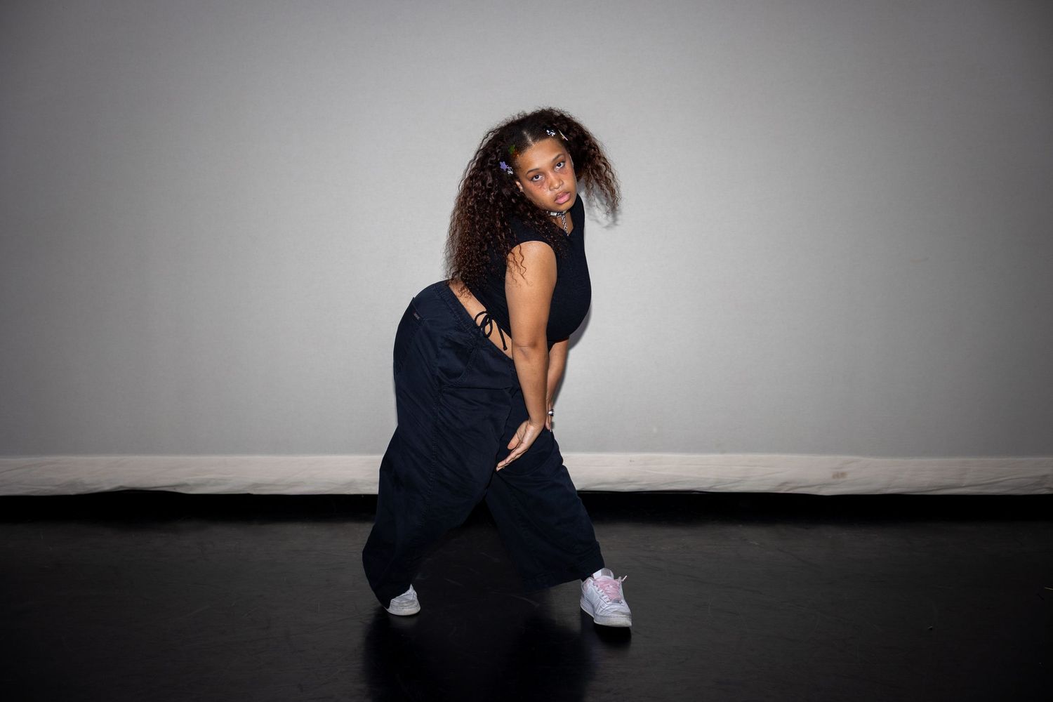 A dancer poses dynamically in black overalls and white sneakers against a gray studio background.