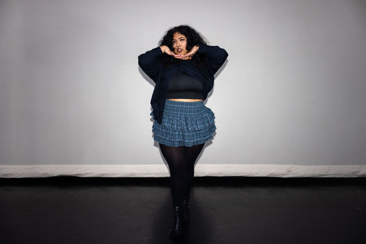 Curvy woman posing in blue plaid mini skirt and black crop top against gray wall.