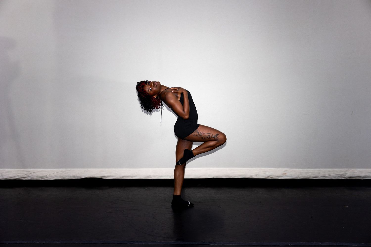 A dancer balances on one leg in a dramatic backbend pose wearing a black bodysuit in a studio.