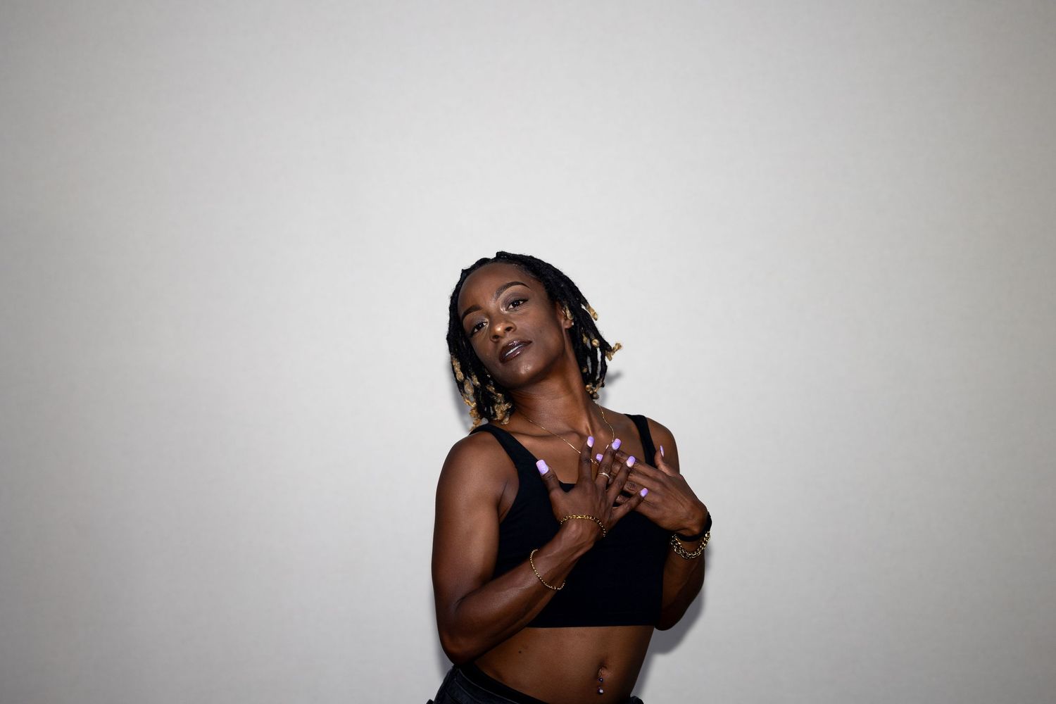 A Black woman in a black sports bra clasps her hands to her chest while posing against a white wall.