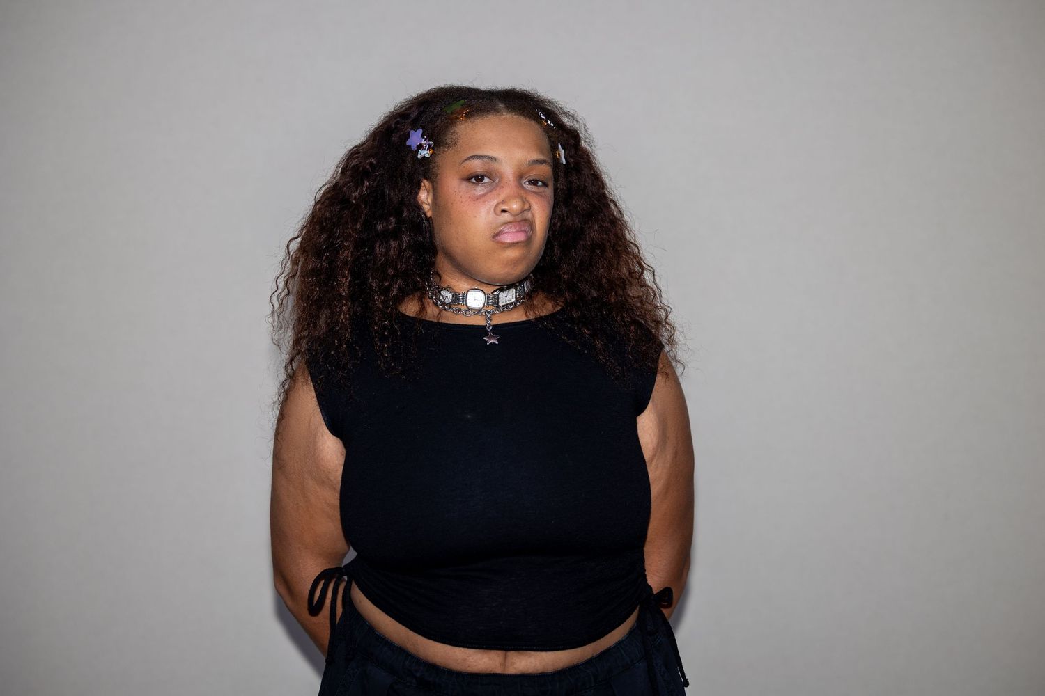A curly-haired woman in a black sleeveless crop top and silver choker necklace poses against a gray background.