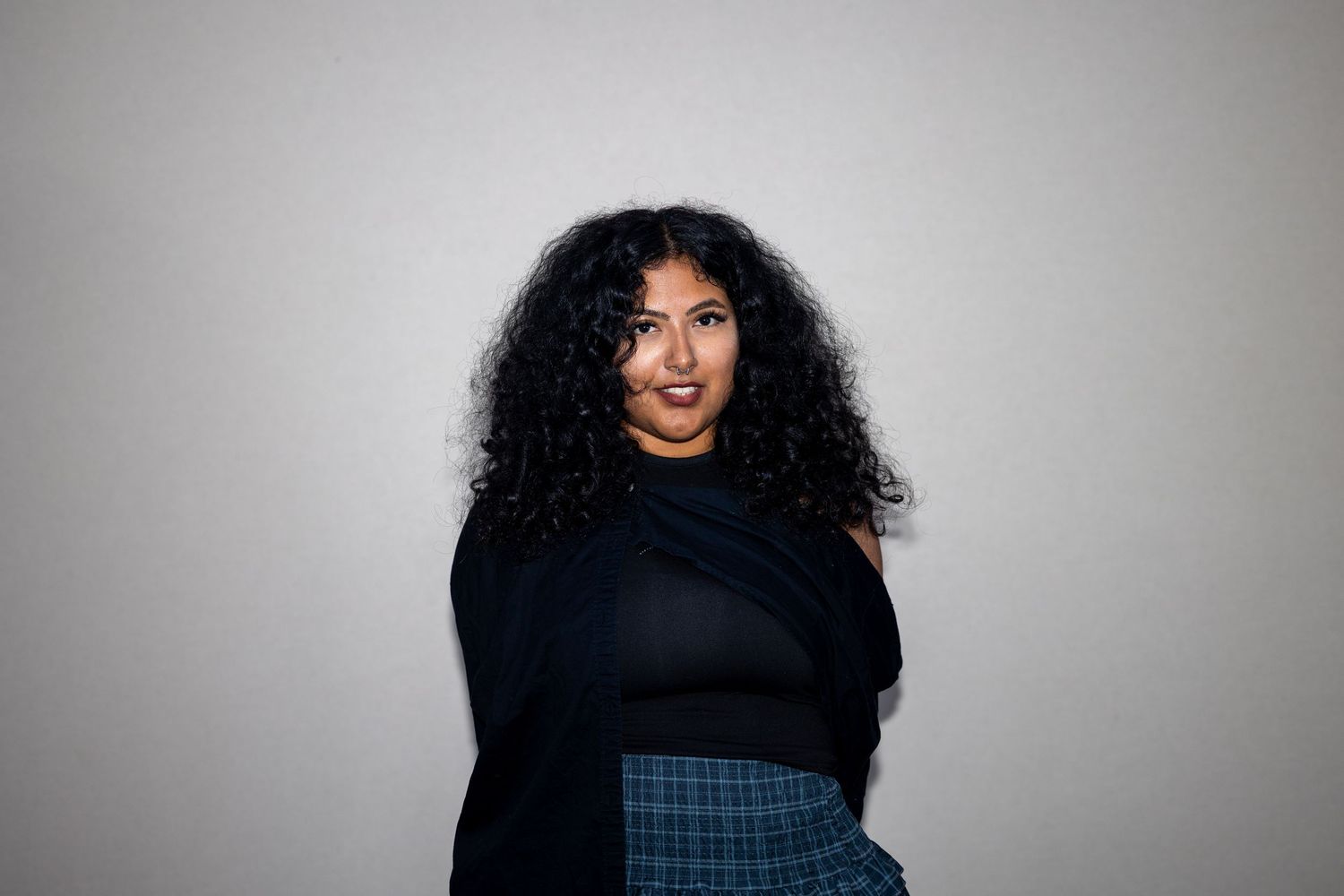 A plus-size woman with voluminous curly hair wears a black wrap top and plaid skirt against a gray background.