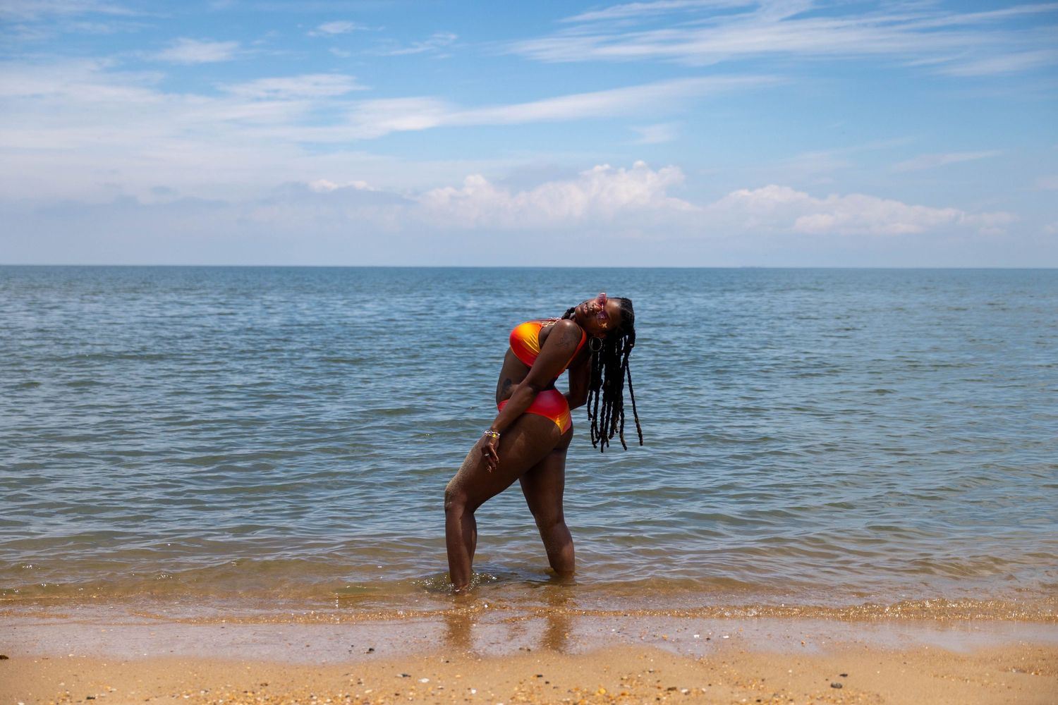 Woman in red and yellow bikini dancing on sandy beach with ocean and cloudy sky behind her.
