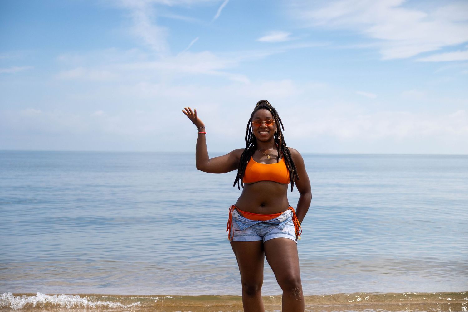 A joyful young Black woman in an orange crop top and denim shorts poses happily on a sunny beach by the ocean.