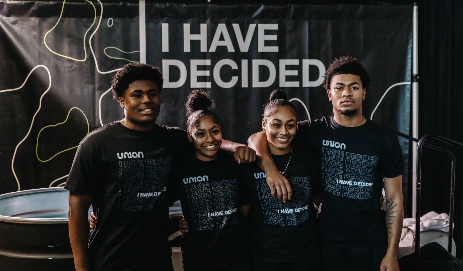 A group of athletes stand together in a gym wearing dark colored shirts with text.