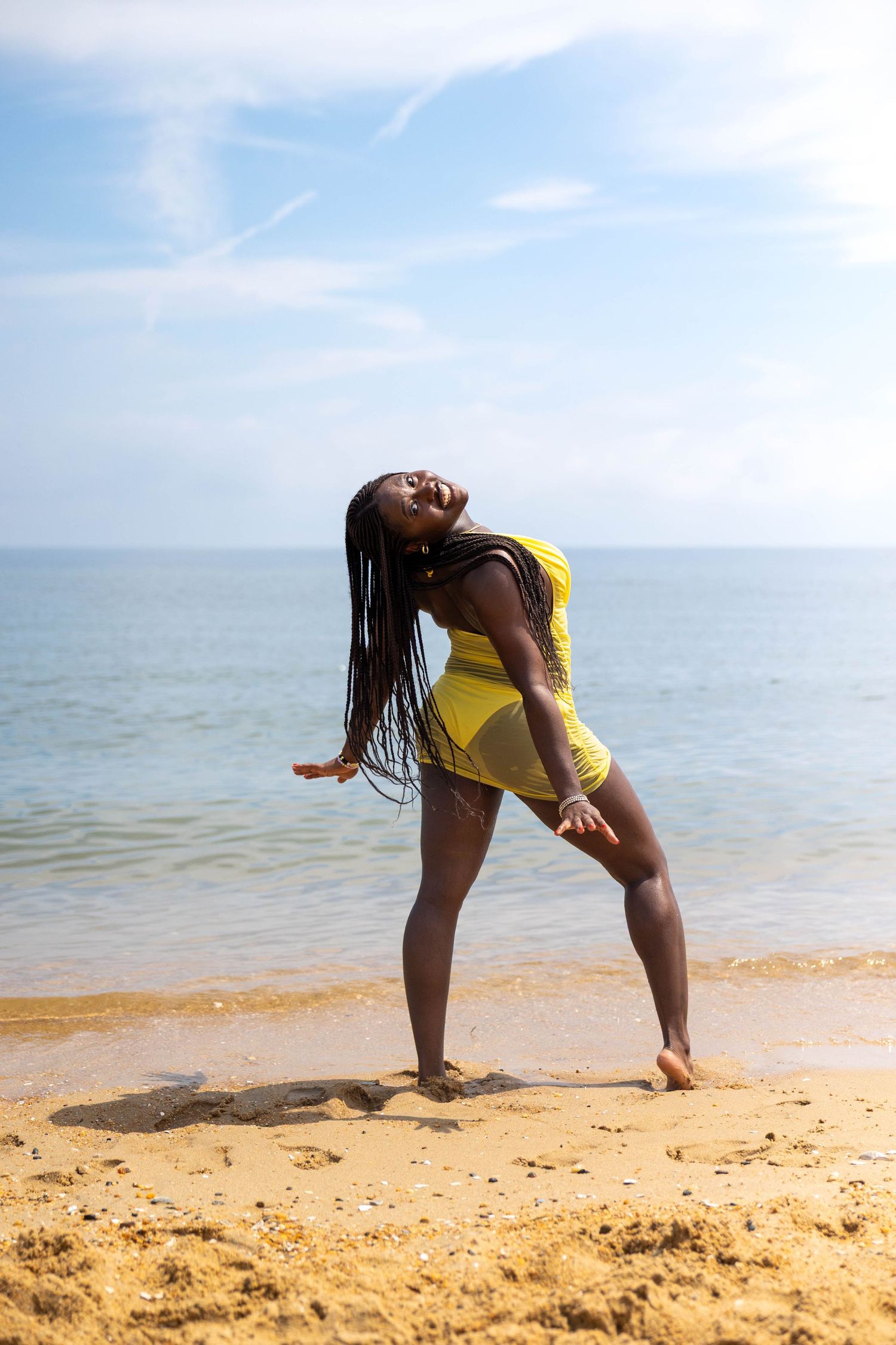 A joyful Black woman in a yellow swimsuit strikes a dynamic pose on a sandy beach with blue sky and ocean in the background.