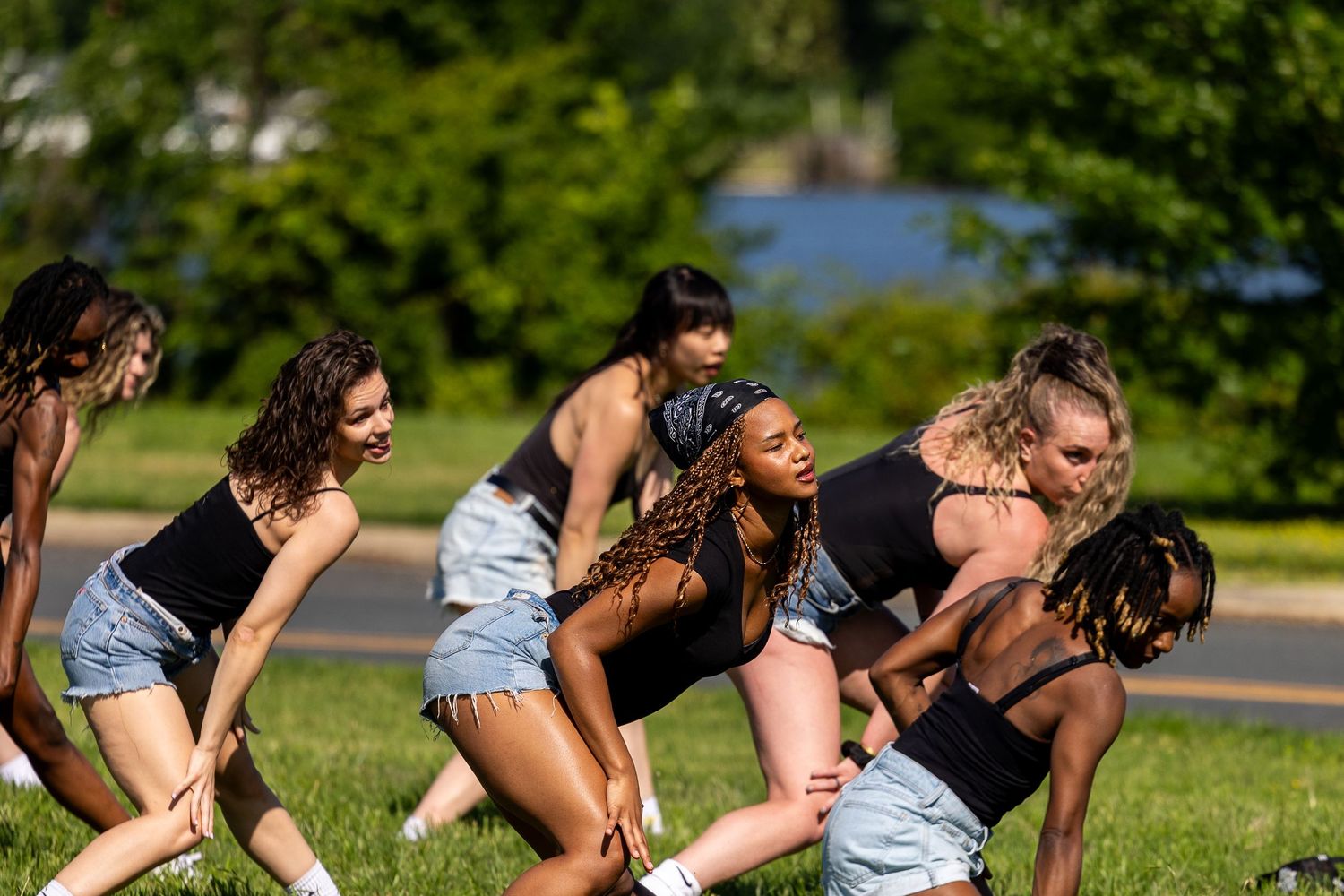 A group of young women in black tops and denim shorts perform a dance routine outdoors on a sunny day.