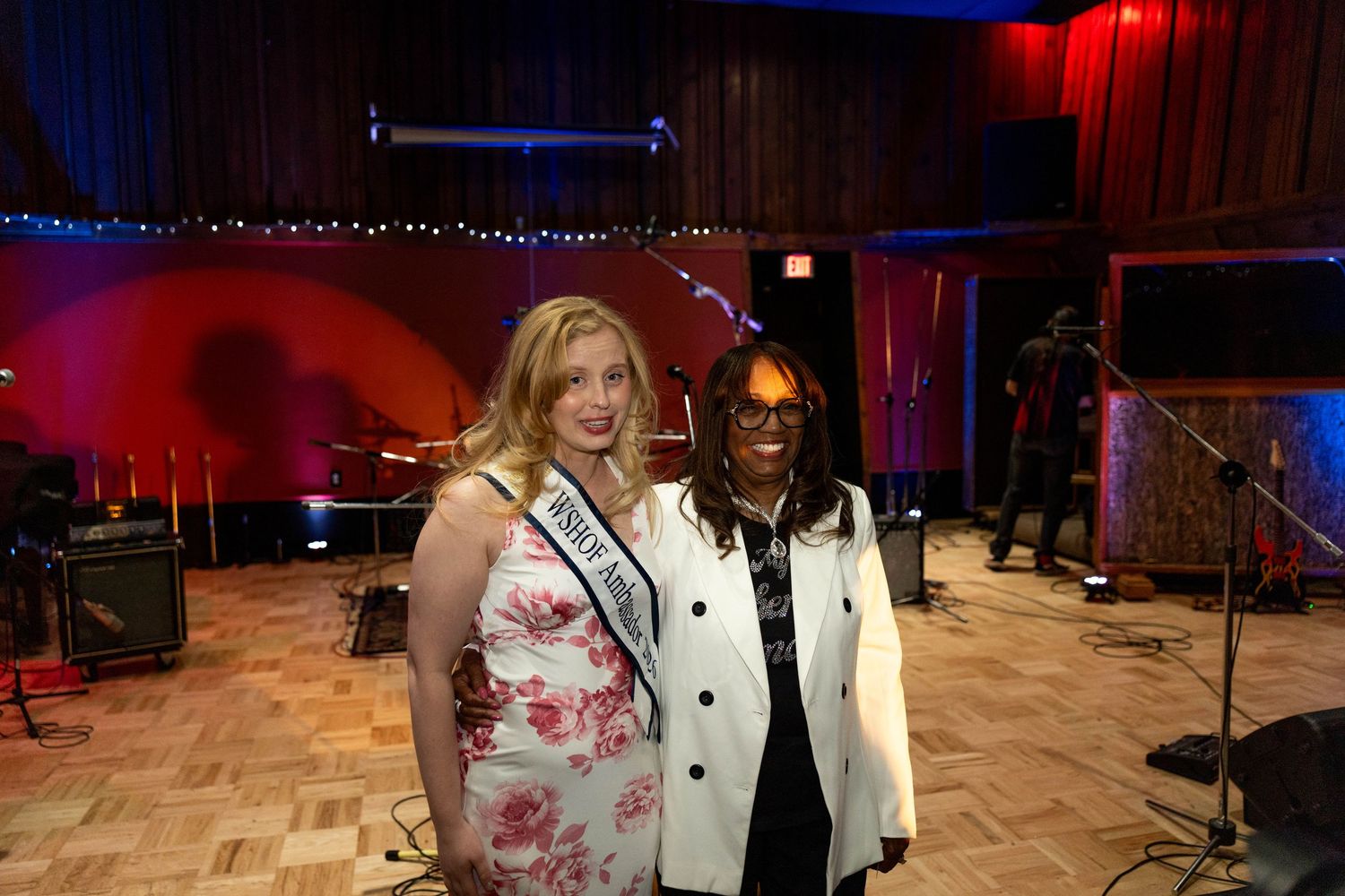 Two women posing together at an indoor venue, one wearing a floral dress with a pageant sash.