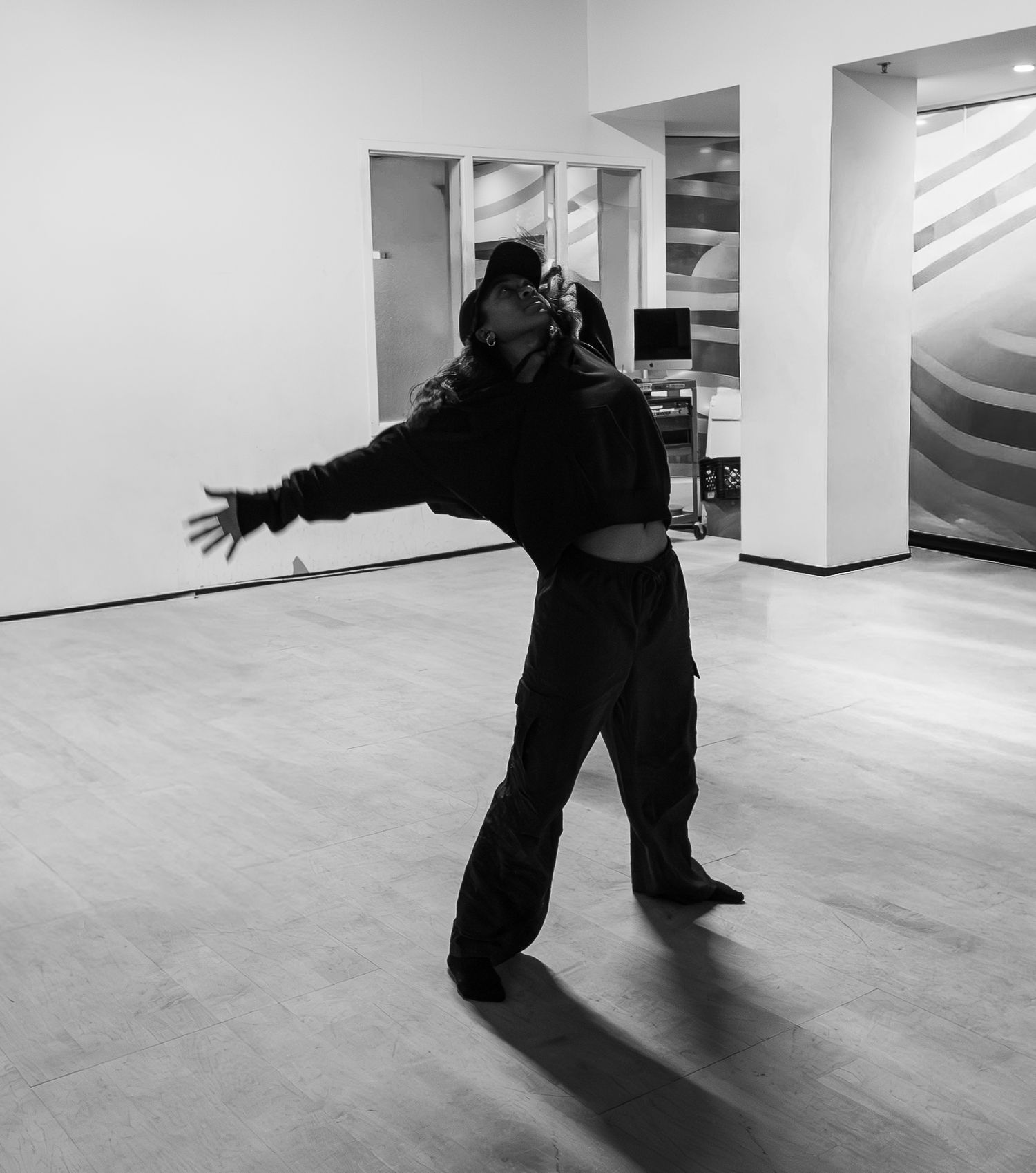 A dancer in all-black outfit strikes an expressive pose with arms outstretched in a modern dance studio.